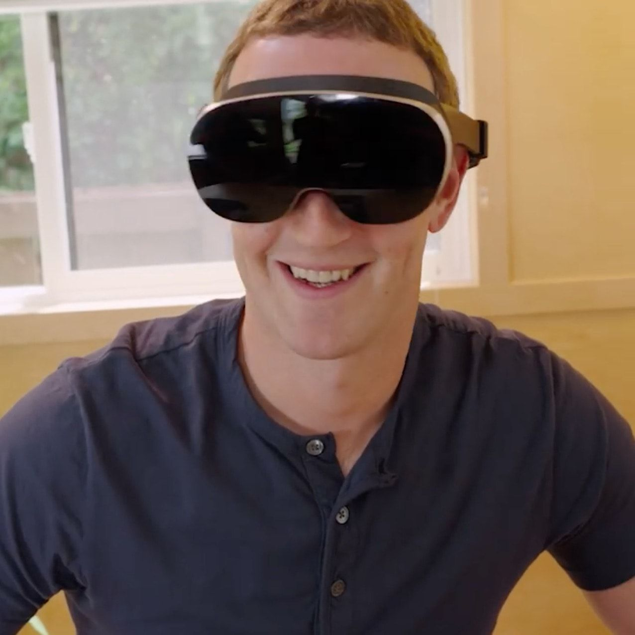Homem branco, sorridente, com camisa azul escuro com botões, usando um headset de realidade virtual com a frente preta, brilhante.