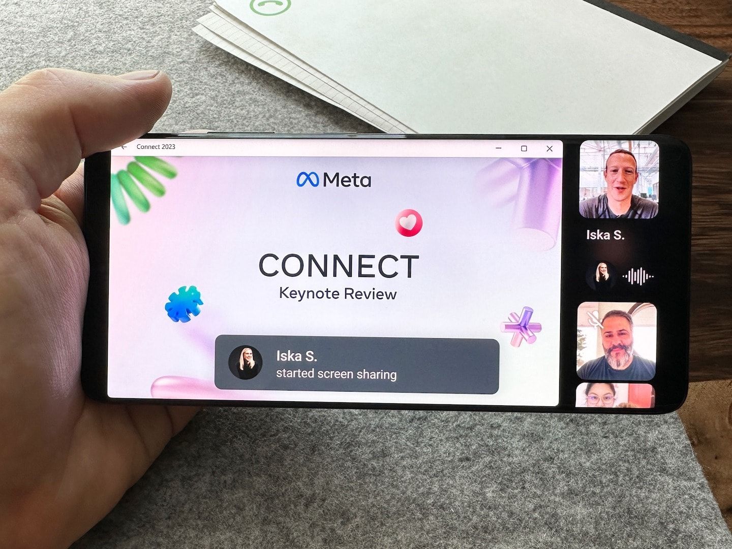 Foto de um celular segurado por uma mão, na horizontal, exibindo uma tela compartilhada durante videochamada no WhatsApp.